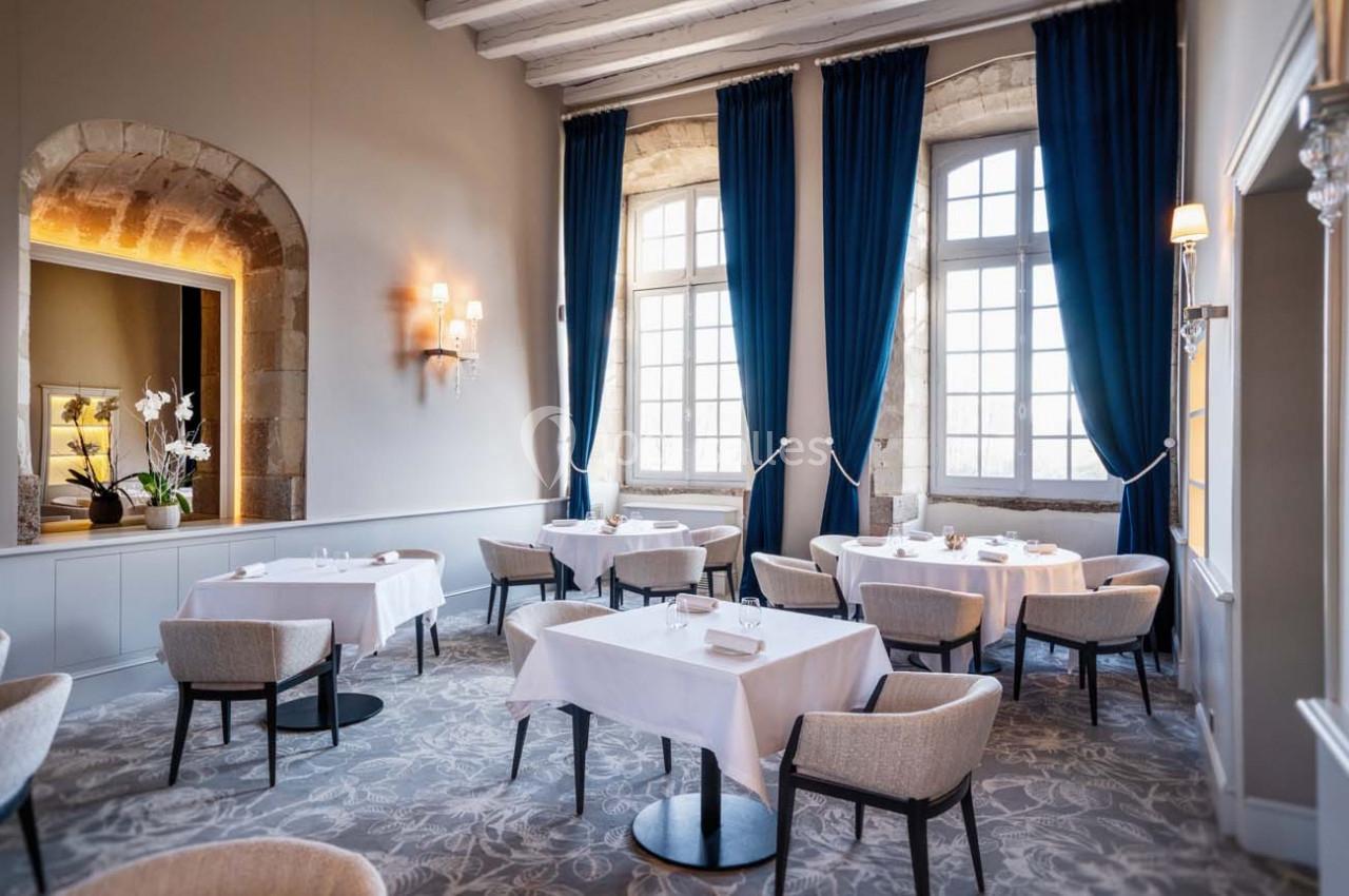 Salle de restaurant élégante avec tables dressées, grandes fenêtres encadrées de rideaux bleus et murs en pierre apparente.