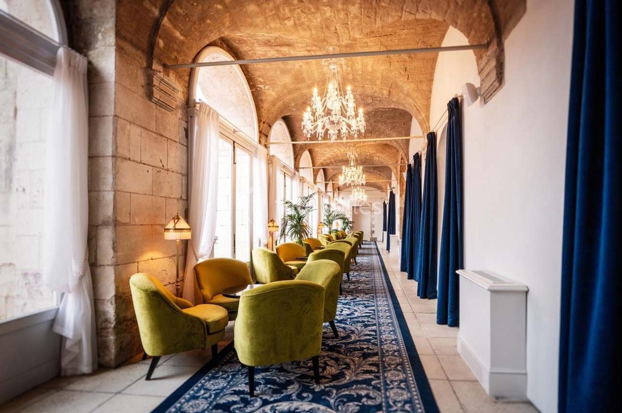 Couloir lumineux avec fauteuils verts, tapis bleu, rideaux foncés et lustres suspendus sous des voûtes en pierre.