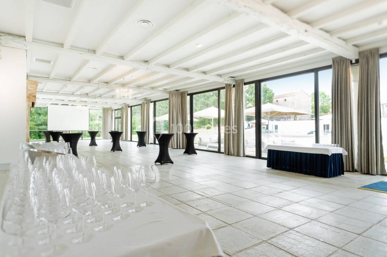 Salle lumineuse avec grandes baies vitrées, tables hautes noires, rangée de verres et vue sur une terrasse extérieure.