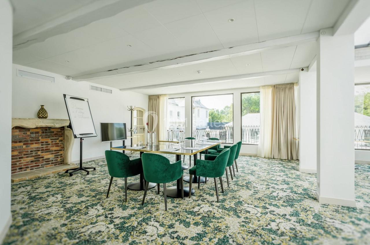 Salle de réunion lumineuse avec table, chaises vertes, tableau blanc, écran et grandes baies vitrées donnant sur l'extérieur.