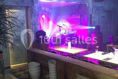 Salle sombre avec éclairage violet, plafond métallique, tables et chaises dispersées sur un sol à carreaux.