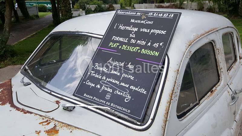 Ardoise posée sur le capot d'une voiture ancienne, affichant un menu de restaurant avec plat, dessert et boisson.