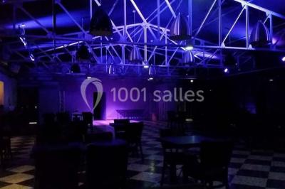 Salle sombre avec éclairage violet, plafond métallique, tables et chaises dispersées sur un sol à carreaux.