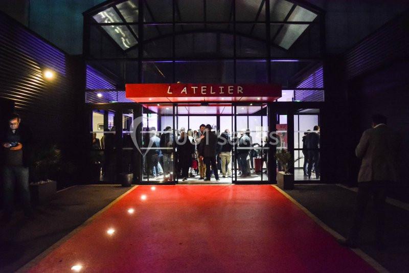 Entrée d'un bâtiment nommé ’L'Atelier’ avec des personnes rassemblées à l'intérieur, vue depuis un tapis rouge éclairé.