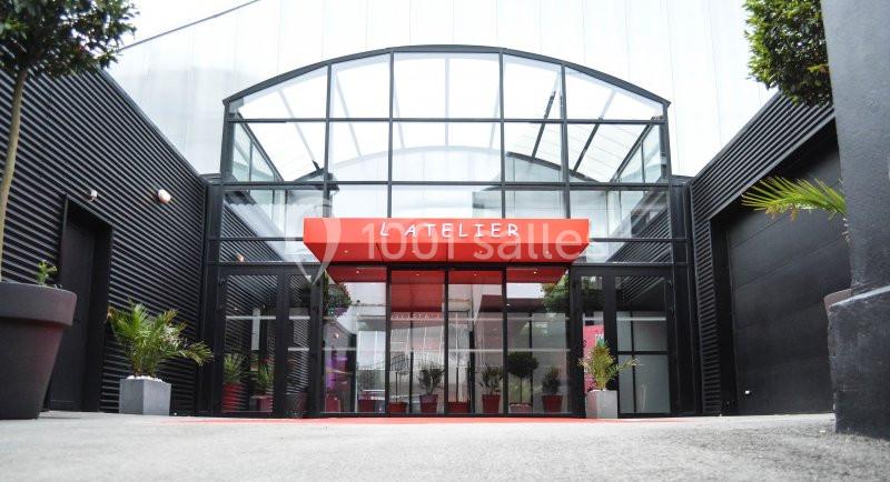 Entrée vitrée moderne d'un bâtiment avec une enseigne rouge indiquant ’L'Atelier’, entourée de plantes en pots.