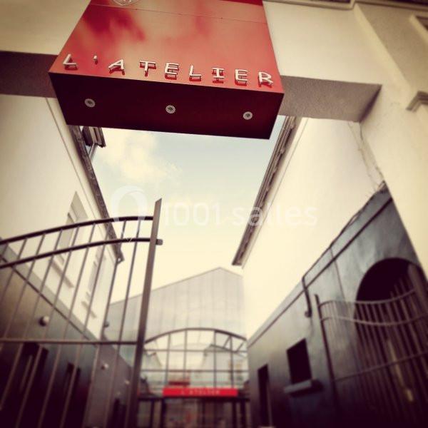 Enseigne rouge indiquant ’L'Atelier’ au-dessus d'une entrée avec portail métallique et bâtiments modernes en arrière-plan.