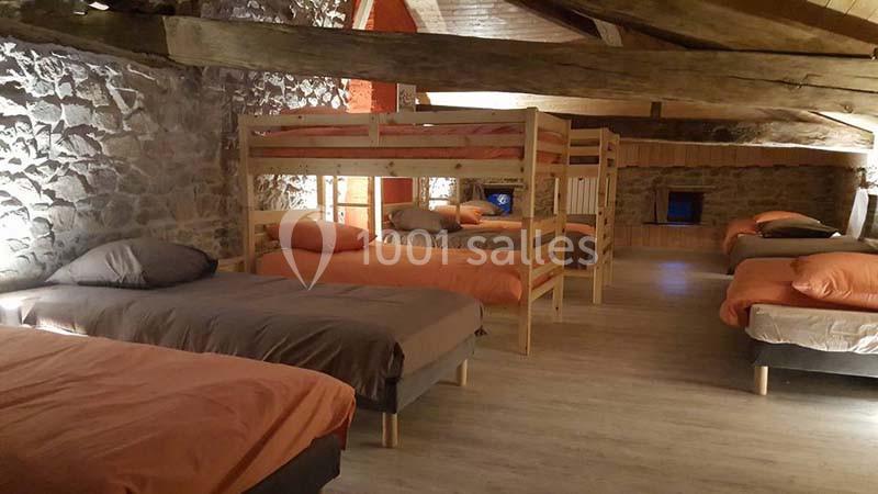 Dortoir avec lits simples et superposés, draps orange et gris, dans une pièce aux murs en pierre et poutres apparentes.