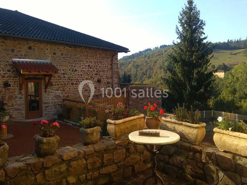 Cour en pierre avec des jardinières fleuries, table ronde et vue sur un bâtiment en pierre et un paysage vallonné boisé.