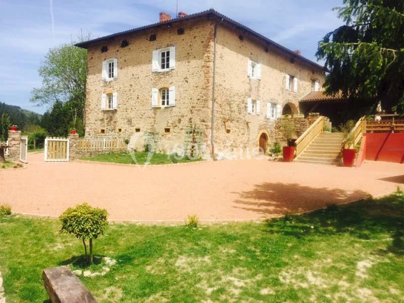 Grande maison en pierre avec volets blancs, entourée d'une cour gravillonnée et d'un jardin verdoyant.