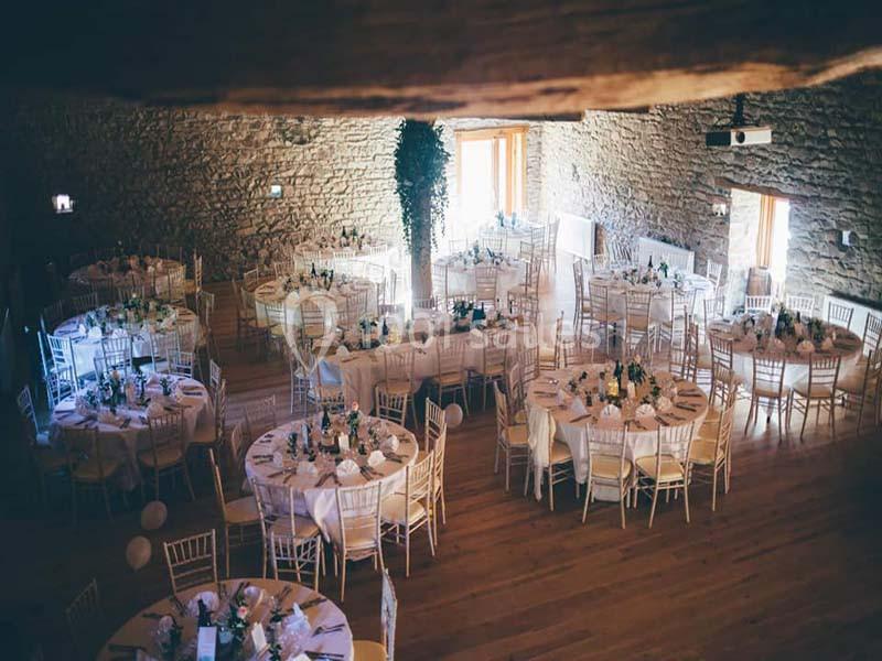 Salle en pierre avec des tables rondes décorées pour un événement, éclairée par la lumière naturelle.