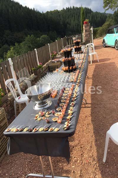 Table dressée en extérieur avec des amuse-bouches, des verres et une grande coupe en métal, près d'une clôture en bois.