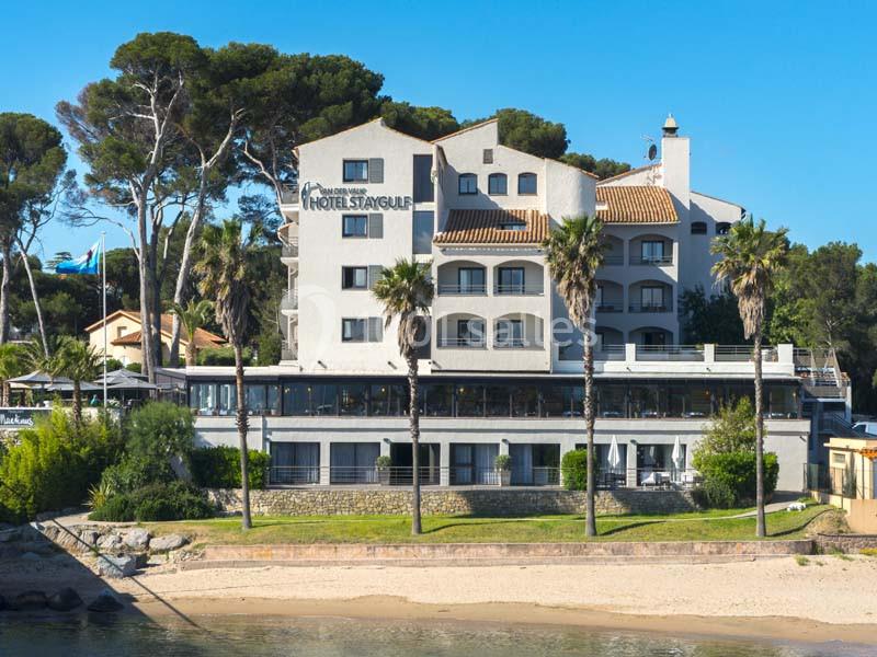 Façade d'un hôtel en bord de mer, entouré de palmiers et de pins, avec une plage au premier plan.
