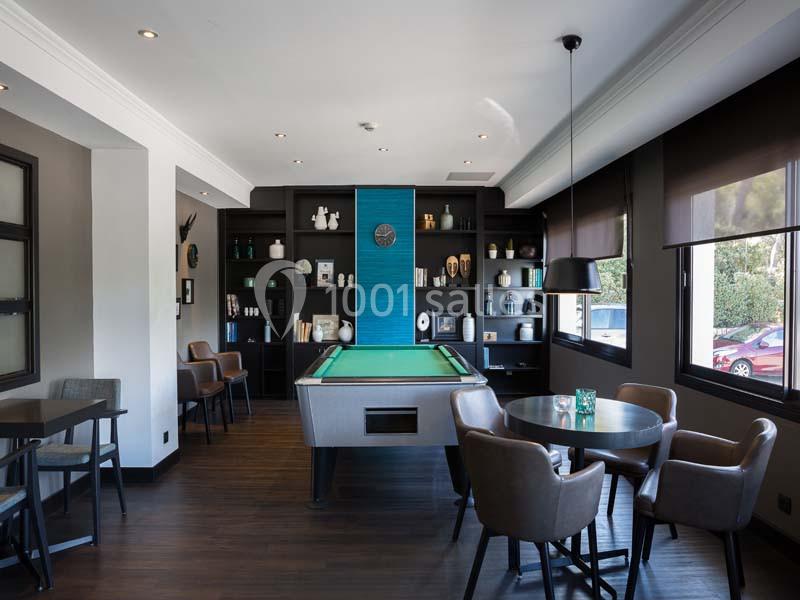 Salle moderne avec table de billard centrale, étagères décoratives, tables et chaises, éclairée par des fenêtres.