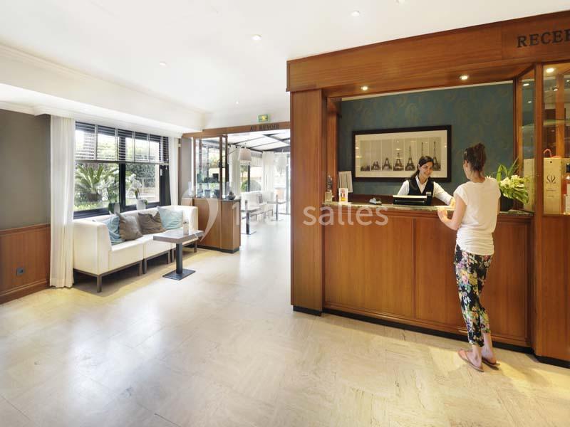 Une femme échange avec une réceptionniste dans le hall lumineux d'un hôtel, avec un coin salon visible en arrière-plan.