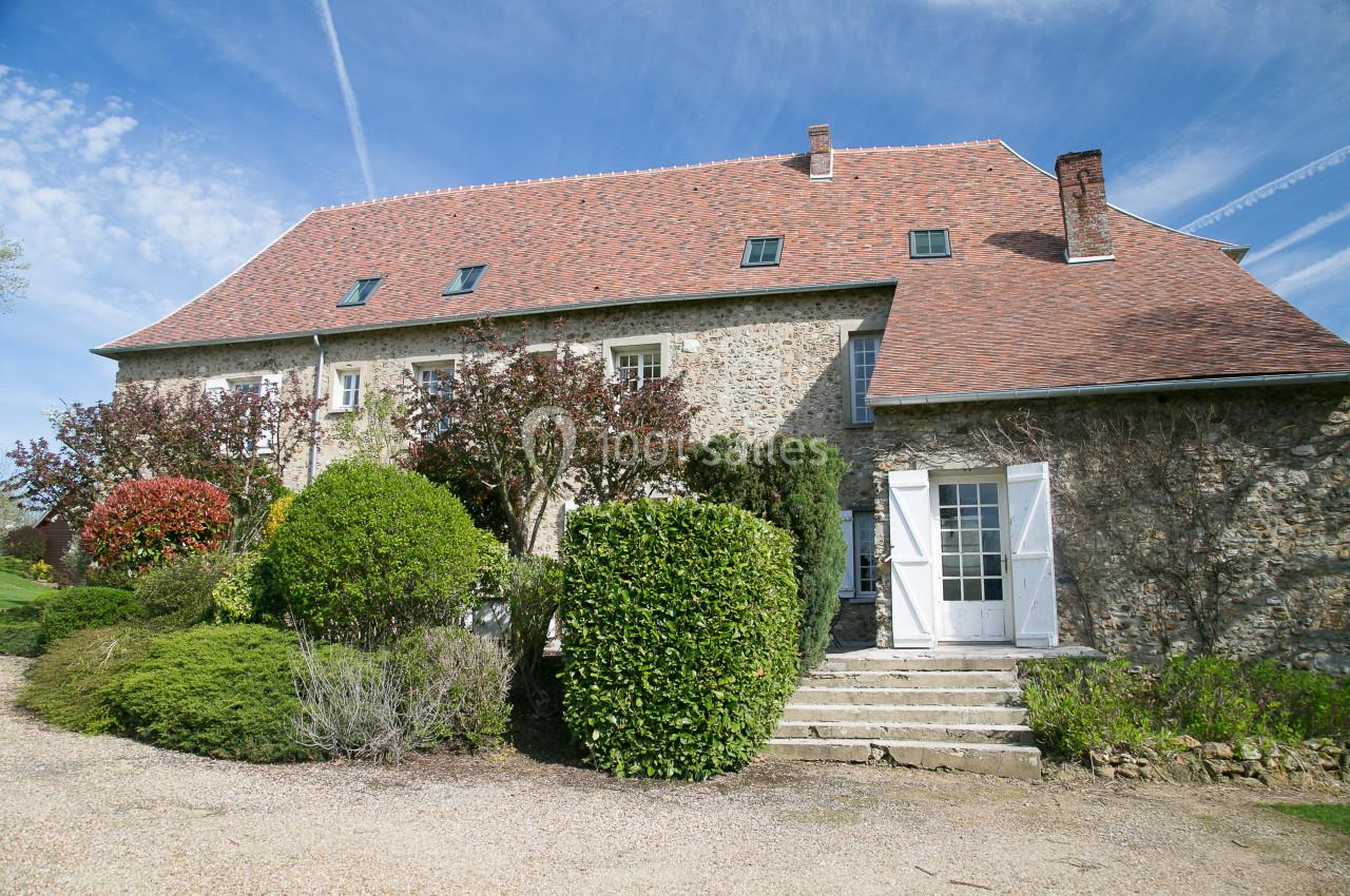 Façade d'une maison en pierre avec toit en tuiles, entourée de buissons et d'arbres sous un ciel dégagé.