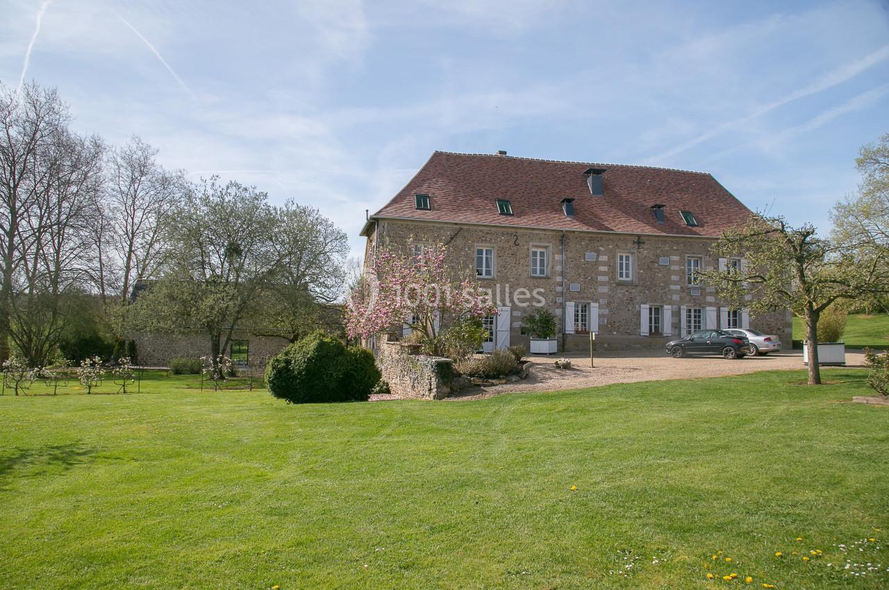 Grande maison en pierre avec toit en tuiles rouges, entourée d'un jardin verdoyant et d'arbres sous un ciel bleu.