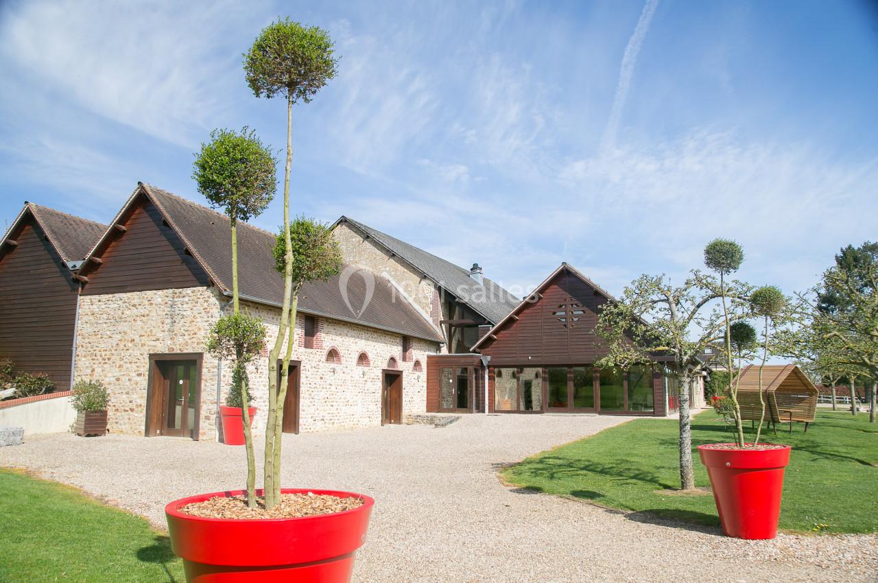 Bâtiment en pierre et bois avec jardin aménagé, arbres taillés et pots rouges sur une allée gravillonnée.