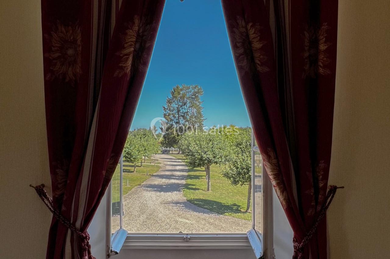 Vue d'un chemin bordé d'arbres à travers une fenêtre encadrée de rideaux rouges ornés de motifs floraux.