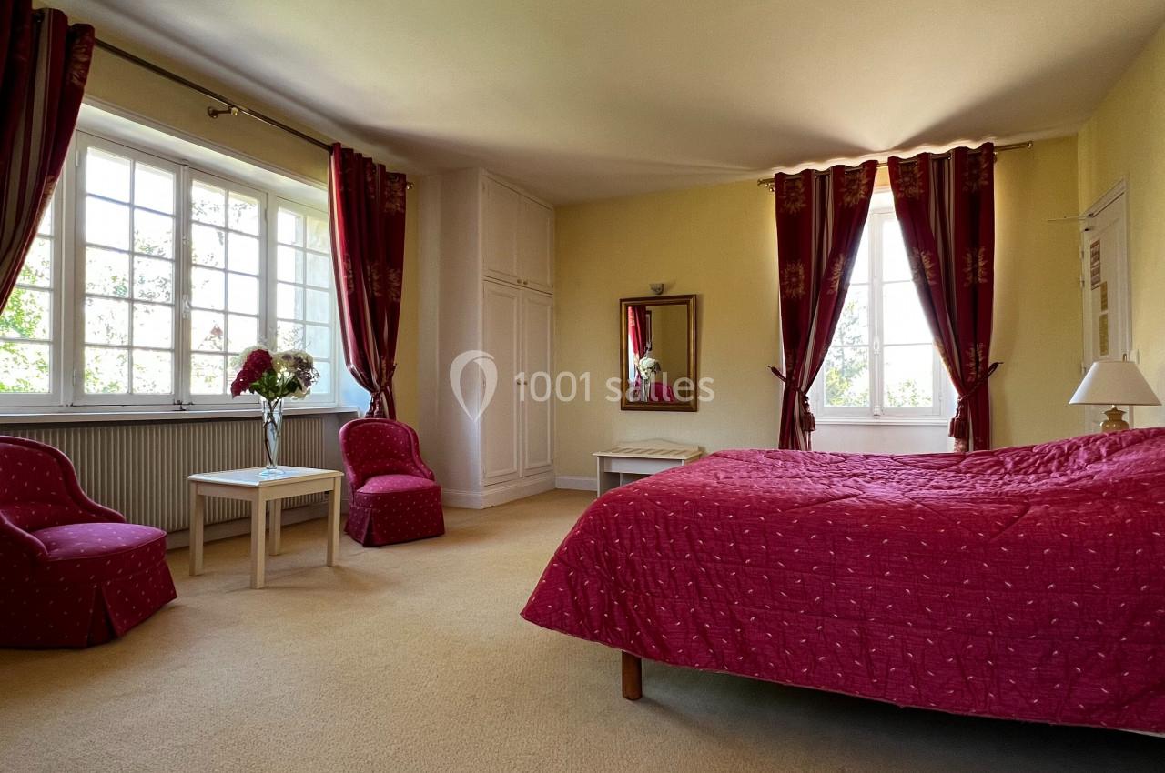 Chambre lumineuse avec lit double, fauteuils rouges, tables basses, grandes fenêtres et rideaux assortis.