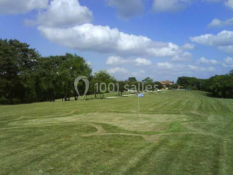 Terrain de golf verdoyant avec un drapeau au premier plan, entouré d'arbres sous un ciel partiellement nuageux.