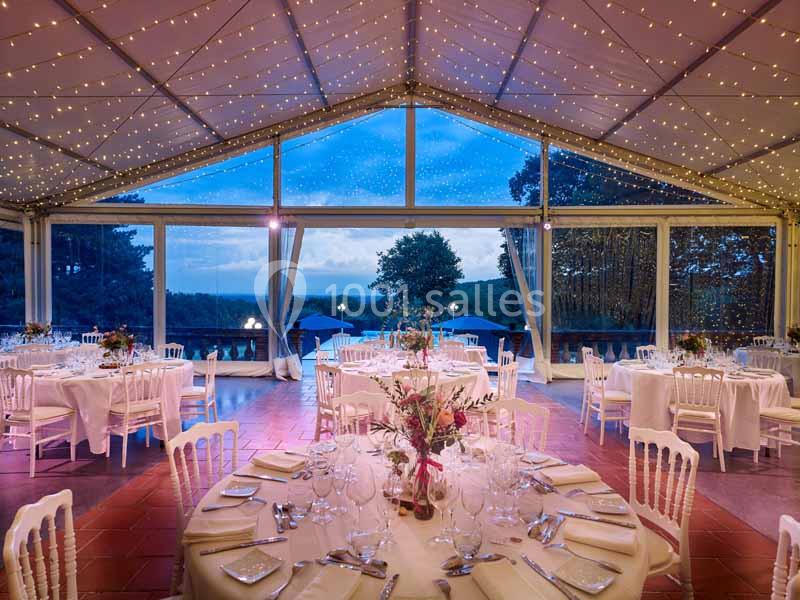 Salle de réception sous une tente éclairée de guirlandes lumineuses, avec des tables dressées et vue sur un paysage…