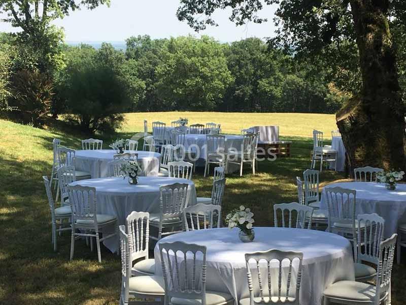 Tables rondes dressées avec nappes blanches et chaises, disposées en extérieur sous des arbres dans un cadre verdoyant.