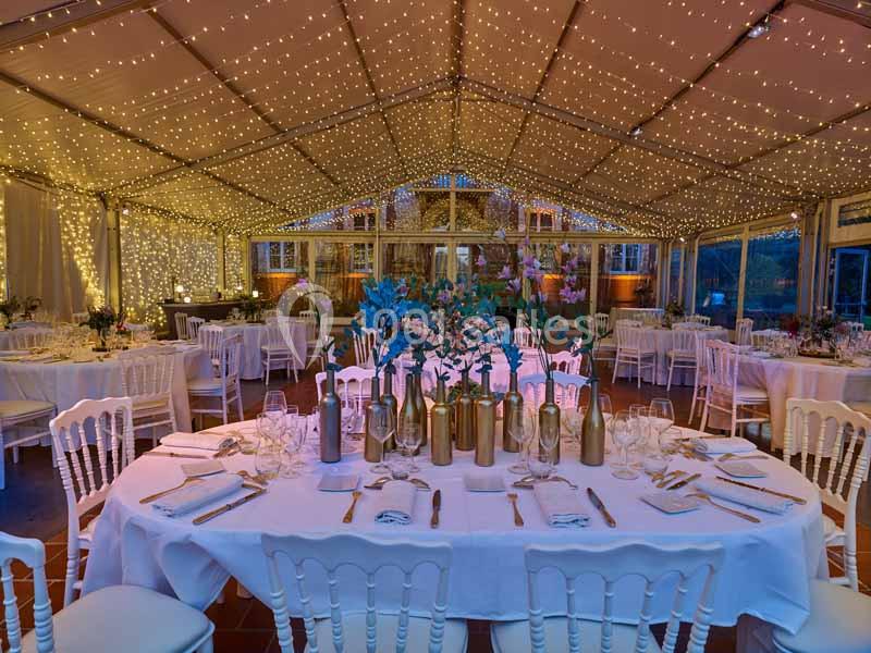 Salle de réception décorée avec des guirlandes lumineuses, tables rondes dressées et vases fleuris au centre.