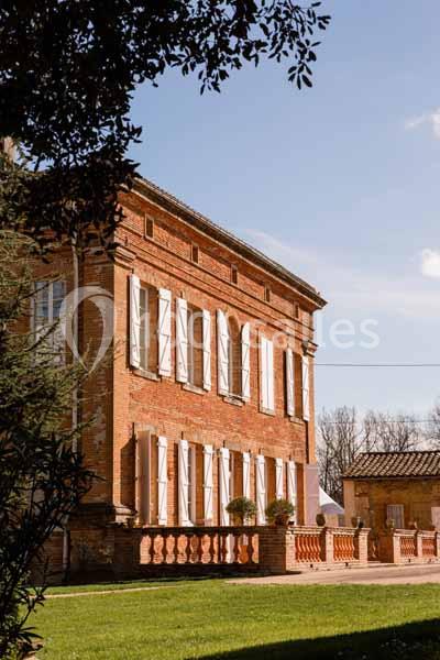 Façade d'une maison en briques rouges avec volets blancs, entourée de verdure sous un ciel dégagé.