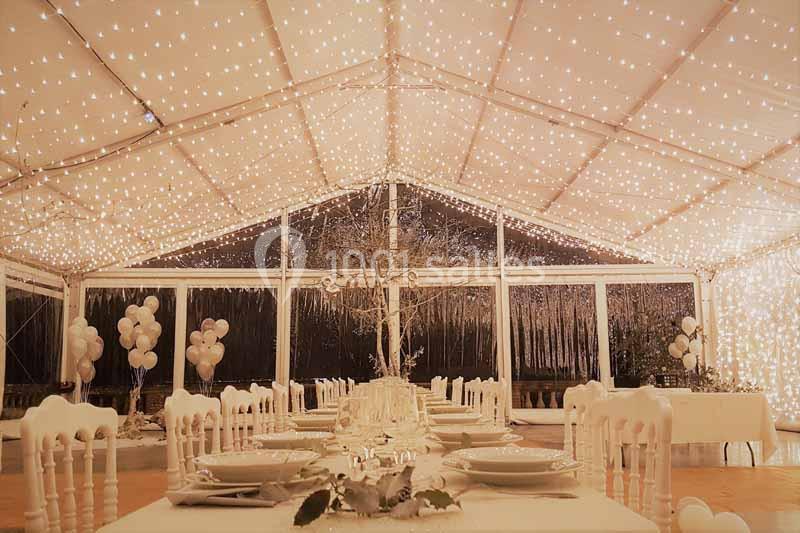 Salle de réception décorée avec des guirlandes lumineuses, tables dressées et ballons blancs en arrière-plan.