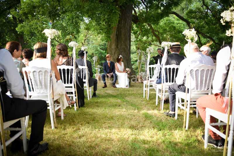 Cérémonie de mariage en plein air avec des invités assis sur des chaises blanches, sous des arbres verdoyants.
