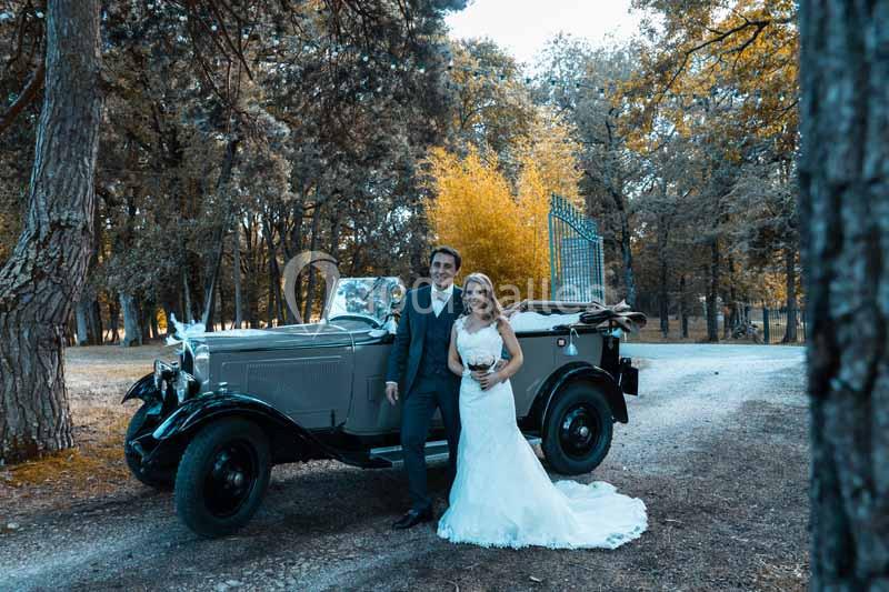 Un couple en tenue de mariage pose devant une voiture ancienne dans un cadre boisé.