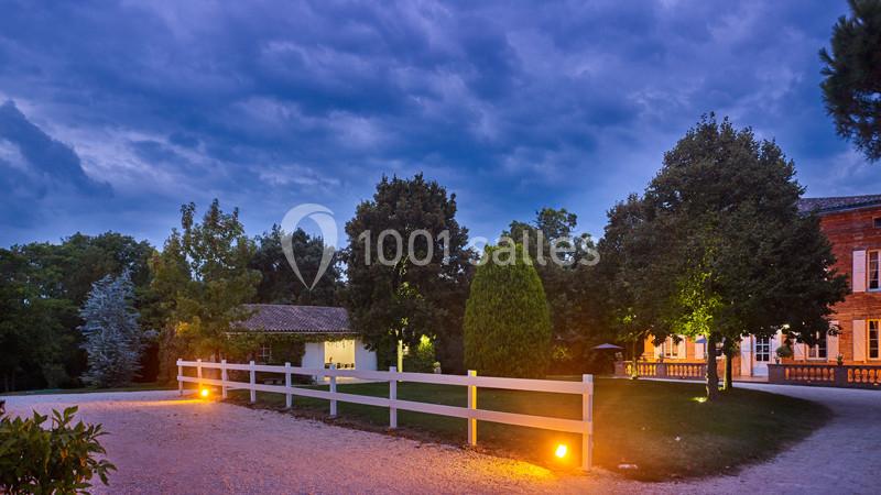 Cour extérieure d'une propriété au crépuscule, éclairée par des lampes au sol, entourée d'arbres et de bâtiments.
