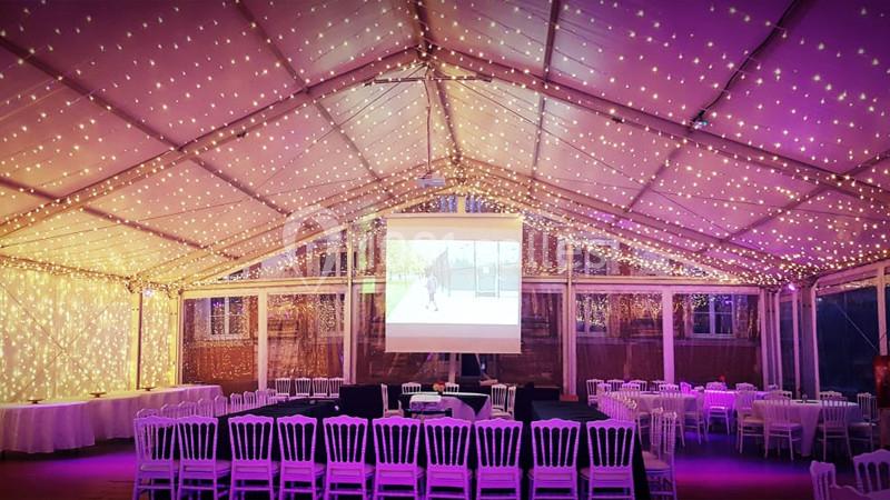 Salle de réception sous une tente illuminée de guirlandes lumineuses, avec des tables et des chaises blanches disposées.