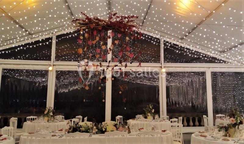 Salle de réception sous une tente illuminée, décorée de guirlandes lumineuses et d'une suspension florale centrale.