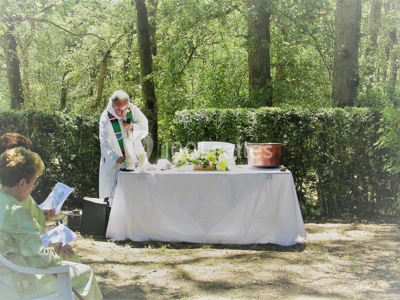 Un prêtre en tenue liturgique officie à une table blanche décorée de fleurs dans un cadre boisé en extérieur.