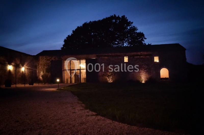 Façade d'un bâtiment en pierre éclairé à la tombée de la nuit, entouré d'une cour gravillonnée et d'arbres.