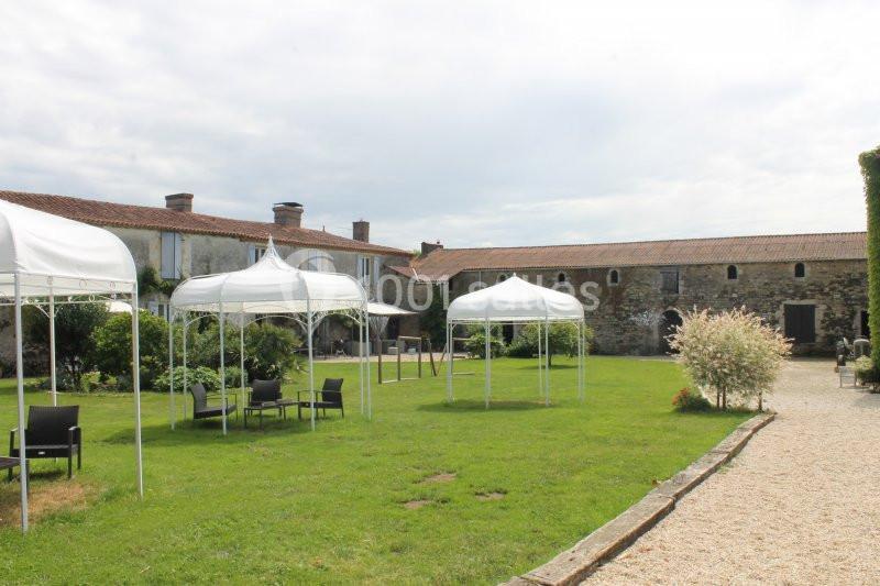 Jardin avec pelouse, tonnelles blanches, mobilier de jardin et bâtiments en pierre sous un ciel partiellement nuageux.