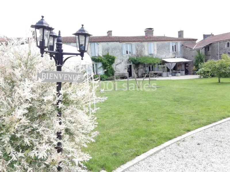 Maison en pierre avec jardin verdoyant, terrasse aménagée et panneau ’Bienvenue’ près d'un lampadaire.