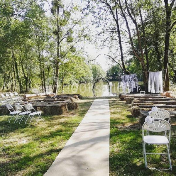 Allée centrale menant à une cérémonie en plein air, entourée de chaises blanches et de bottes de foin près d'un plan d'eau.