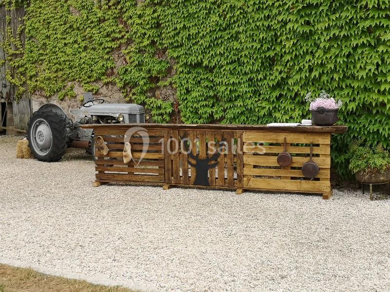 Comptoir en palettes décoré d'ustensiles, installé sur du gravier devant un mur couvert de lierre et un vieux tracteur.