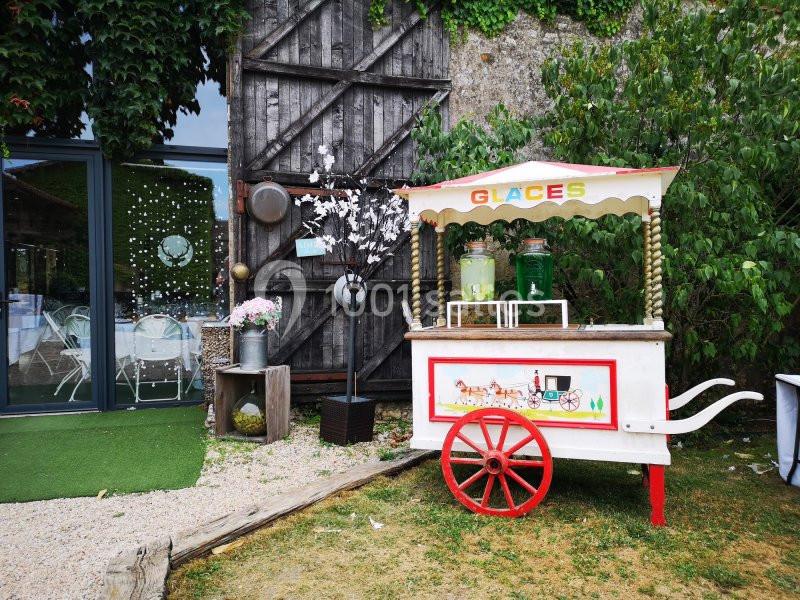Chariot de glaces vintage blanc et rouge devant un mur en bois, entouré de végétation et d'éléments décoratifs.