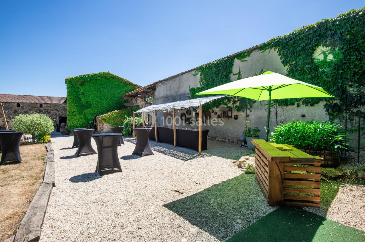 Cour extérieure avec tables hautes noires, parasol vert, végétation et murs en pierre recouverts de lierre.