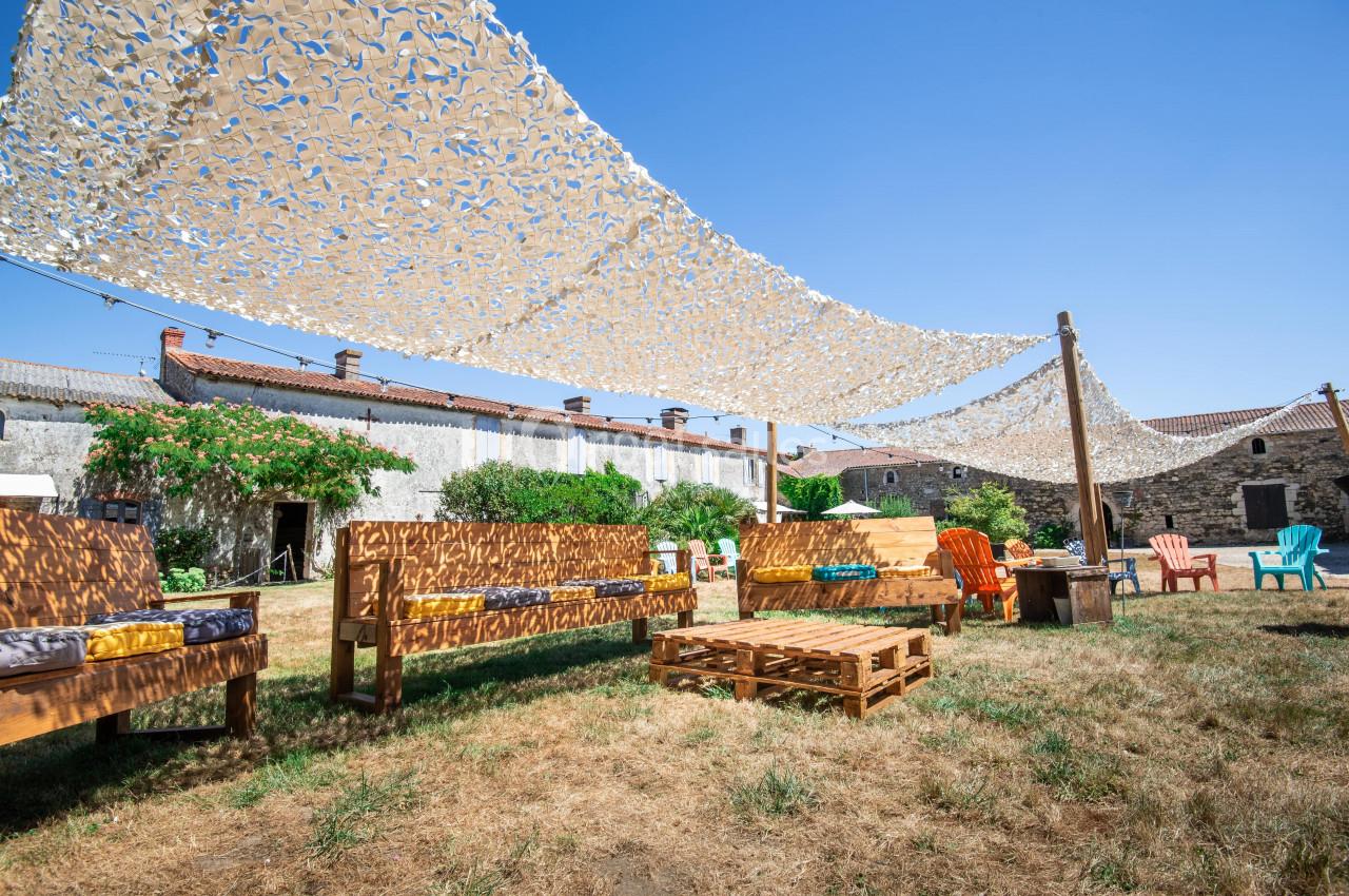 Espace extérieur avec bancs en bois, palettes et chaises sous des voiles d'ombrage, entouré de bâtiments en pierre.