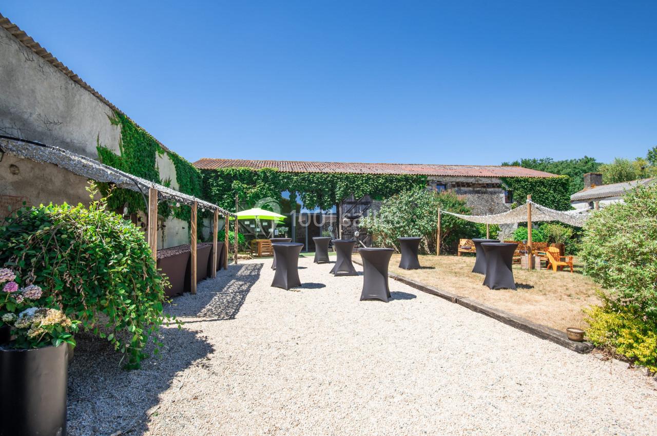 Cour extérieure avec gravier, tables hautes noires, parasols, végétation et bâtiments en pierre sous un ciel bleu.