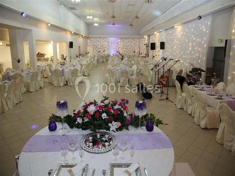 Salle de réception décorée avec des tables rondes, nappes blanches, fleurs et guirlandes lumineuses au mur.