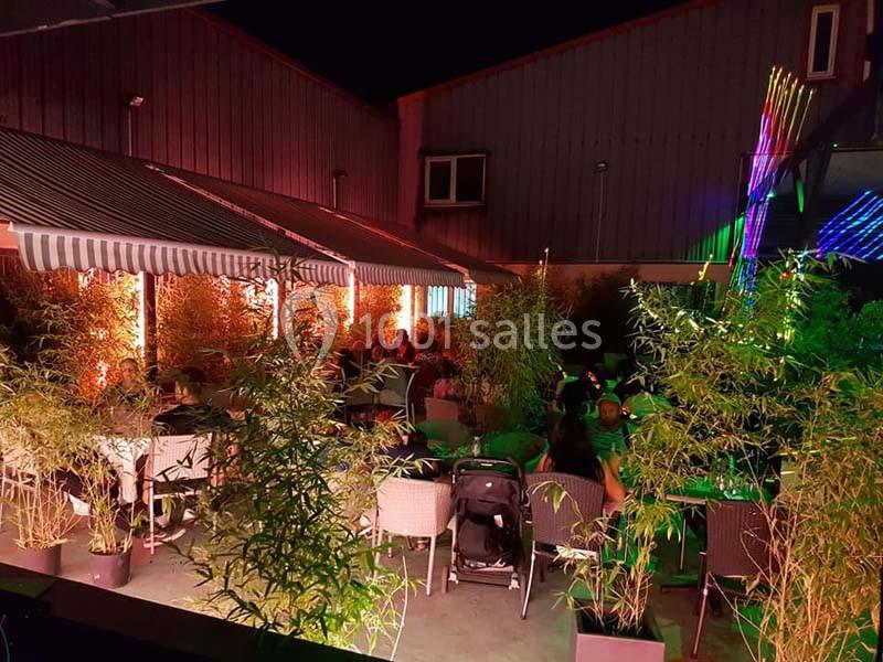 Terrasse extérieure éclairée de nuit, avec des tables entourées de bambous et des lumières colorées.