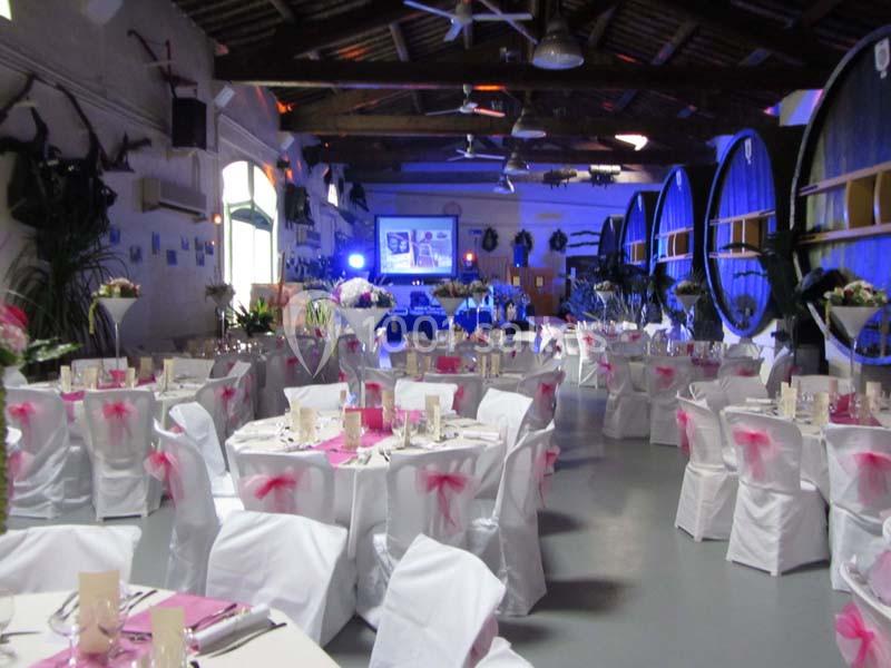Salle décorée pour un événement, avec des tables rondes couvertes de nappes blanches et des chaises ornées de nœuds roses.