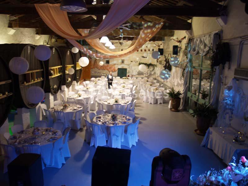 Salle décorée pour un événement, avec des tables rondes dressées, des guirlandes lumineuses et des tonneaux en arrière-plan.