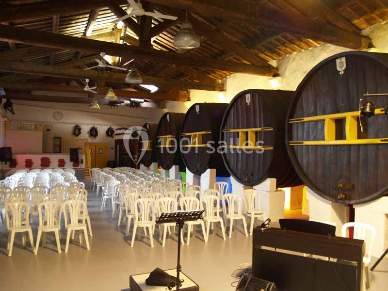 Salle avec des rangées de chaises blanches face à une scène, décorée de grands tonneaux en bois fixés au mur.