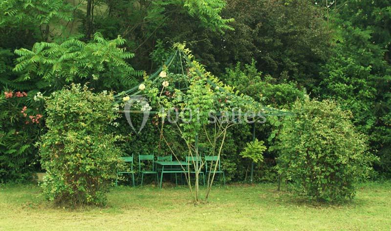 Tonnelle en métal recouverte de végétation, entourée de buissons, avec une table et des chaises vertes en dessous.